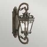 The Tournai Outdoor 4 Light Grand Wall Lantern - Bronze is a vintage-style outdoor fixture, showcasing intricate scrollwork and seedy glass panels within a glass-paneled enclosure. It is mounted on a decorative wall bracket and crafted from metal with an antique bronze finish.