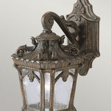 The Tournai Outdoor 1 Light Small Wall Lantern - Bronze radiates a timeless charm with its vintage-style ornate metalwork and intricate scroll designs, enhanced by a rich bronze finish against a plain backdrop.