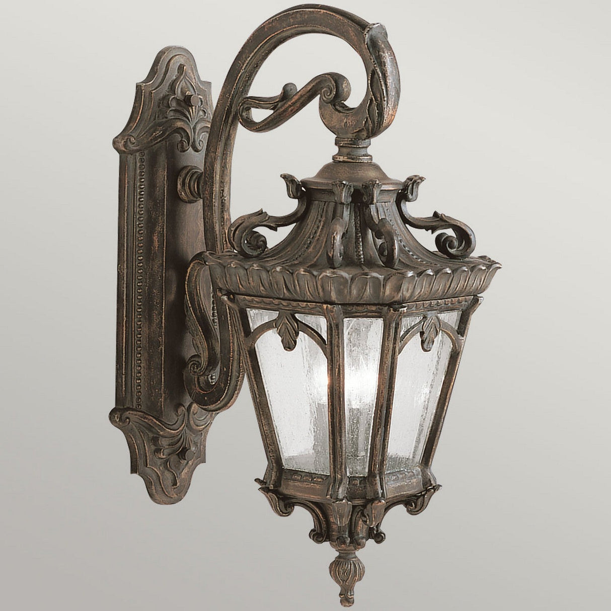 The Tournai Outdoor 3 Light Extra Large Wall Lantern in bronze showcases intricate scrollwork and seedy glass panels, elegantly mounted on a decorative backplate. This design is further enhanced with vintage-inspired detailing.