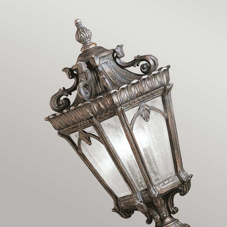 Set against a neutral background, the Tournai 2 Light Large Pedestal in bronze stands proudly with its vintage-style design. This outdoor fixture showcases ornate metalwork and clear seedy glass panels, featuring decorative scrolls and leaf patterns that exude an elegant and classical charm.