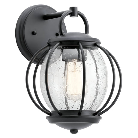 The Vandalia Outdoor 1 Light Wall Lantern - Black features a sleek black frame that encases a clear, seeded glass globe. This elegant fixture highlights the light bulb within and is mounted on a circular base plate, bringing an air of sophistication to your exterior decor.