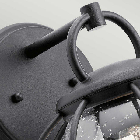 Close-up of the Vandalia Outdoor 1 Light Wall Lantern in black featuring a rounded wall mount. This stylish fixture showcases a curved arm that supports a glass cover with a textured, bubble-like pattern. Set against the light gray background, this piece exemplifies exquisite outdoor lighting design.
