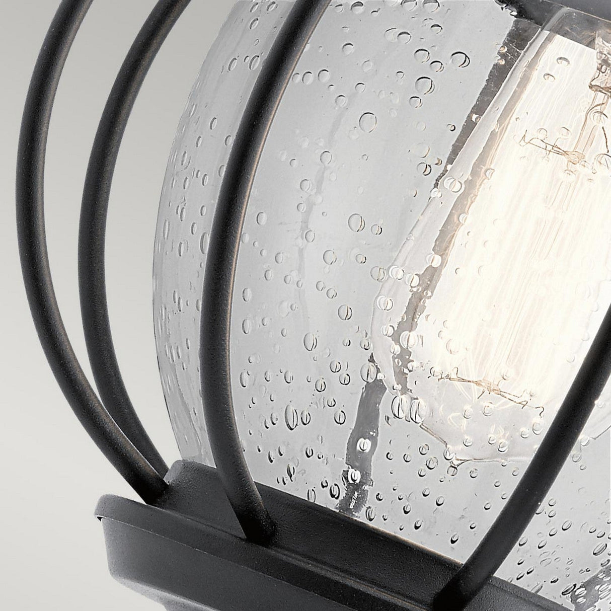 The Vandalia Outdoor 1 Light Wall Lantern in Black showcases a close-up view of its design, featuring a light bulb housed in clear, textured glass surrounded by black metal bars in a cage-like structure. The glass is adorned with speckled water droplets, emphasizing its rustic and industrial appeal for outdoor lighting.