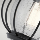 The Vandalia Outdoor 1 Light Wall Lantern in Black showcases a close-up view of its design, featuring a light bulb housed in clear, textured glass surrounded by black metal bars in a cage-like structure. The glass is adorned with speckled water droplets, emphasizing its rustic and industrial appeal for outdoor lighting.