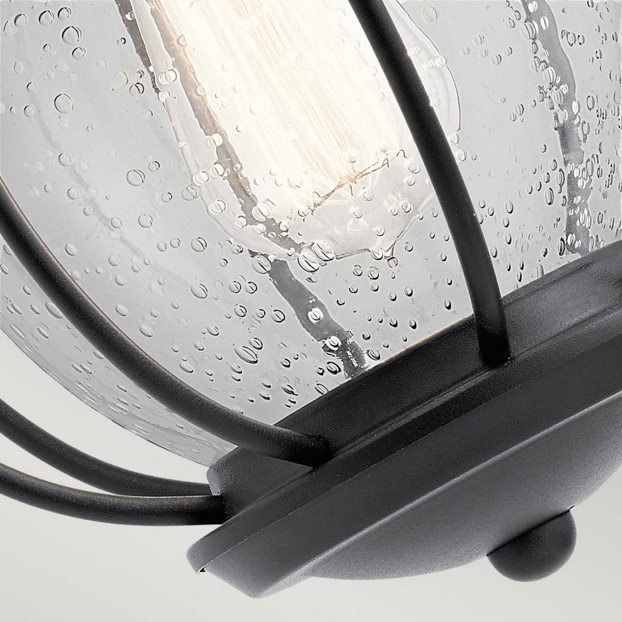 Close-up of the Vandalia Outdoor 1 Light Wall Lantern - Black, featuring raindrop-textured glass. This exterior lighting fixture houses a cylindrical bulb, casting a gentle glow, and is displayed against a simple backdrop.