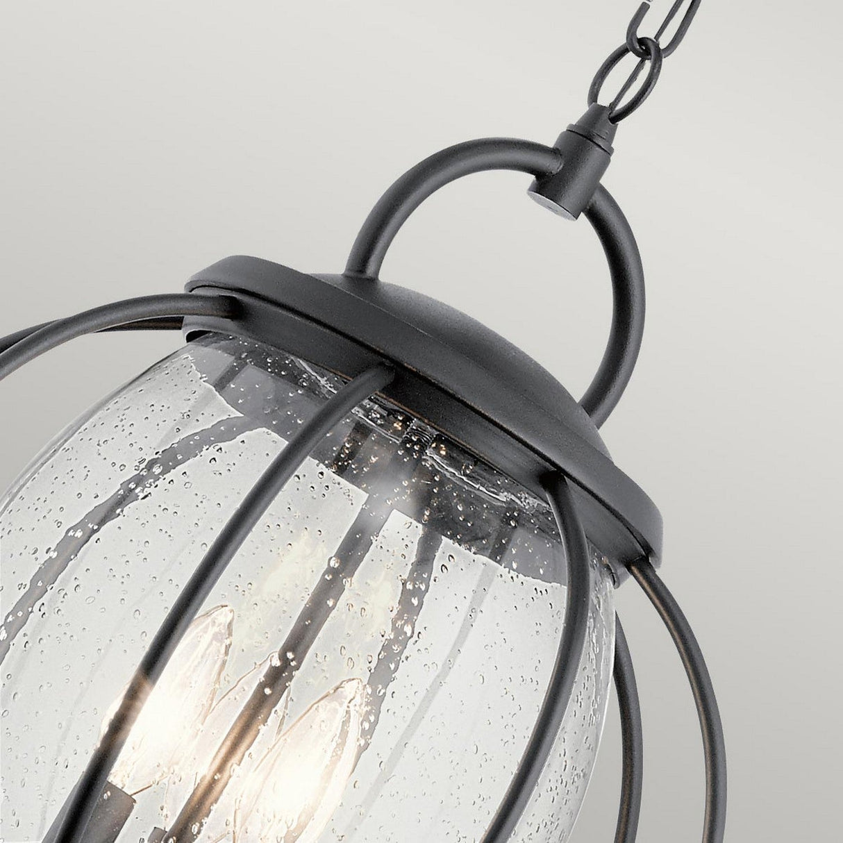 The Vandalia Outdoor 3 Light Large Chain Pendant in black showcases a contemporary lantern design with a hanging light fixture. It features a sleek black metal frame and clear, textured glass cover, housing multiple bulbs that emit a warm glow. This pendant light's chain enhances its elegant appearance, making it ideal for illuminating any space.