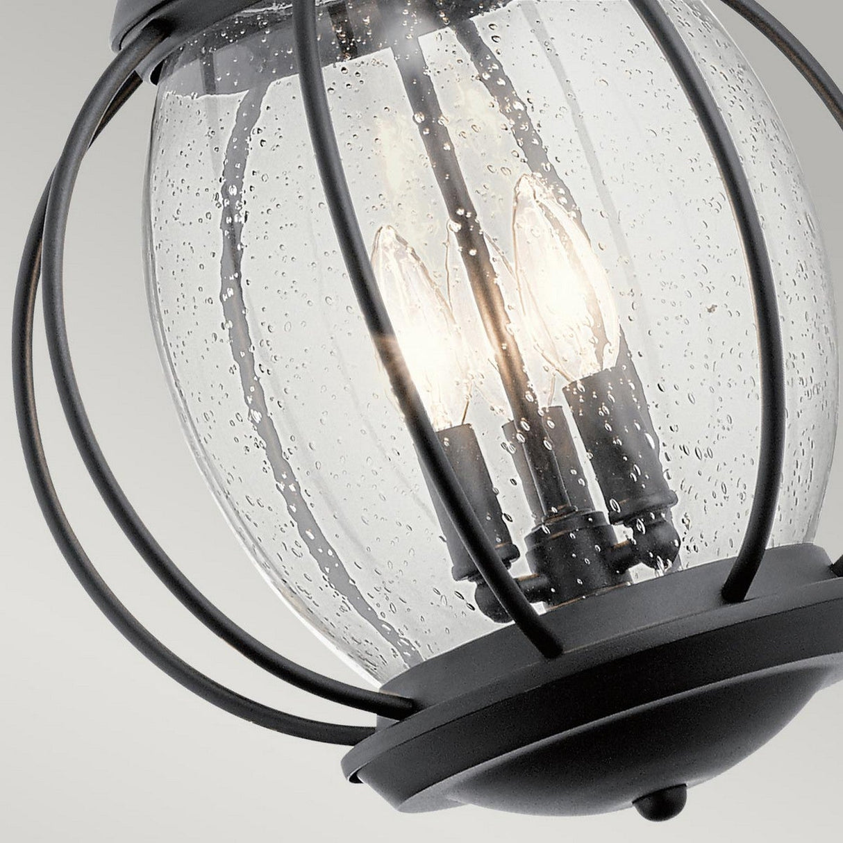A close-up of the Vandalia Outdoor 3 Light Large Chain Pendant in black showcases its modern lantern design with a metal frame and clear bubbled glass, elegantly enclosing three lit bulbs. The curved metal bars contribute to the elegance of this IP44 weatherproof fixture.
