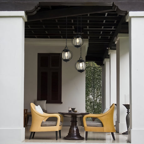 A cozy outdoor seating area features two beige chairs and a round wooden table under a covered patio. The space is illuminated by three Vandalia Outdoor 1 Light Medium Chain Pendants in Black, providing soft lighting. A lush green background enhances the inviting atmosphere, while contemporary design elements complete the look.