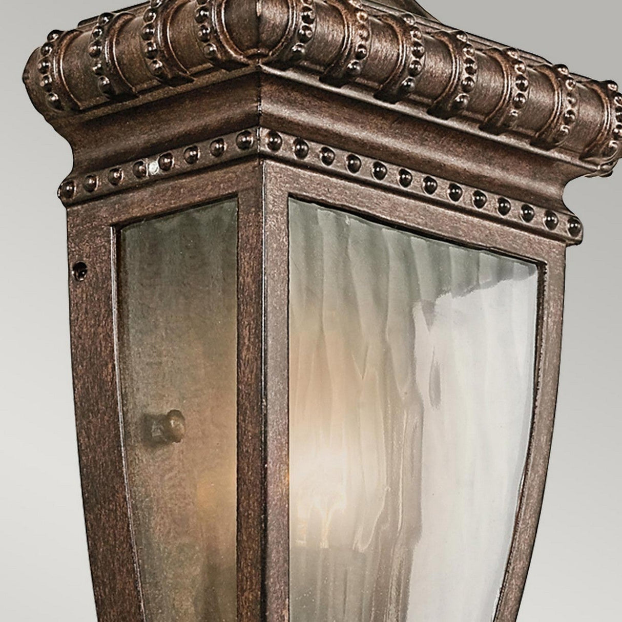 Close-up of the Venetian Rain Outdoor 1 Light Half Wall Lantern in brushed bronze, featuring clear glass panels. The gently glowing light inside accentuates the ornate detailing, evoking a gentle Venetian rain over its vintage-style frame.