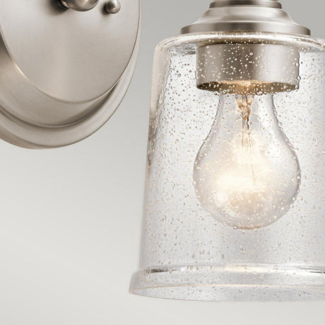 A close-up of the Waverly 1 Light Wall Light - Silver showcases a glowing bulb encased in its clear, textured glass shade, beautifully highlighted by a silver finish that elegantly complements the soft, neutral background.