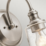 Introducing the Waverly 1 Light Wall Light in Silver: A close-up view reveals its vintage-inspired design, featuring a brushed silver finish and a wall-mounted fixture. The seeded glass shade elegantly showcases a visible light bulb, while the round base and curved arm blend classic elegance with modern flair.