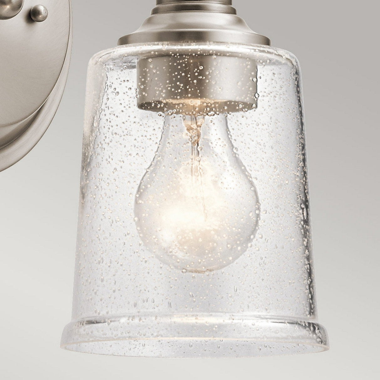 Detailed view of the Waverly 1 Light Wall Light - Silver, showcasing its vintage-inspired design with a clear glass shade adorned with small bubbles. The bulb inside emits a warm glow, and the silver metal fitting offers a modern contrast against the gray background.