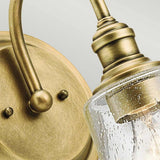 Close-up of the Waverly 1 Light Wall Light in Natural Brass, showcasing a clear, seeded glass shade. The fixture features an elegant curved design with a weathered finish, enhancing its classic and vintage charm.