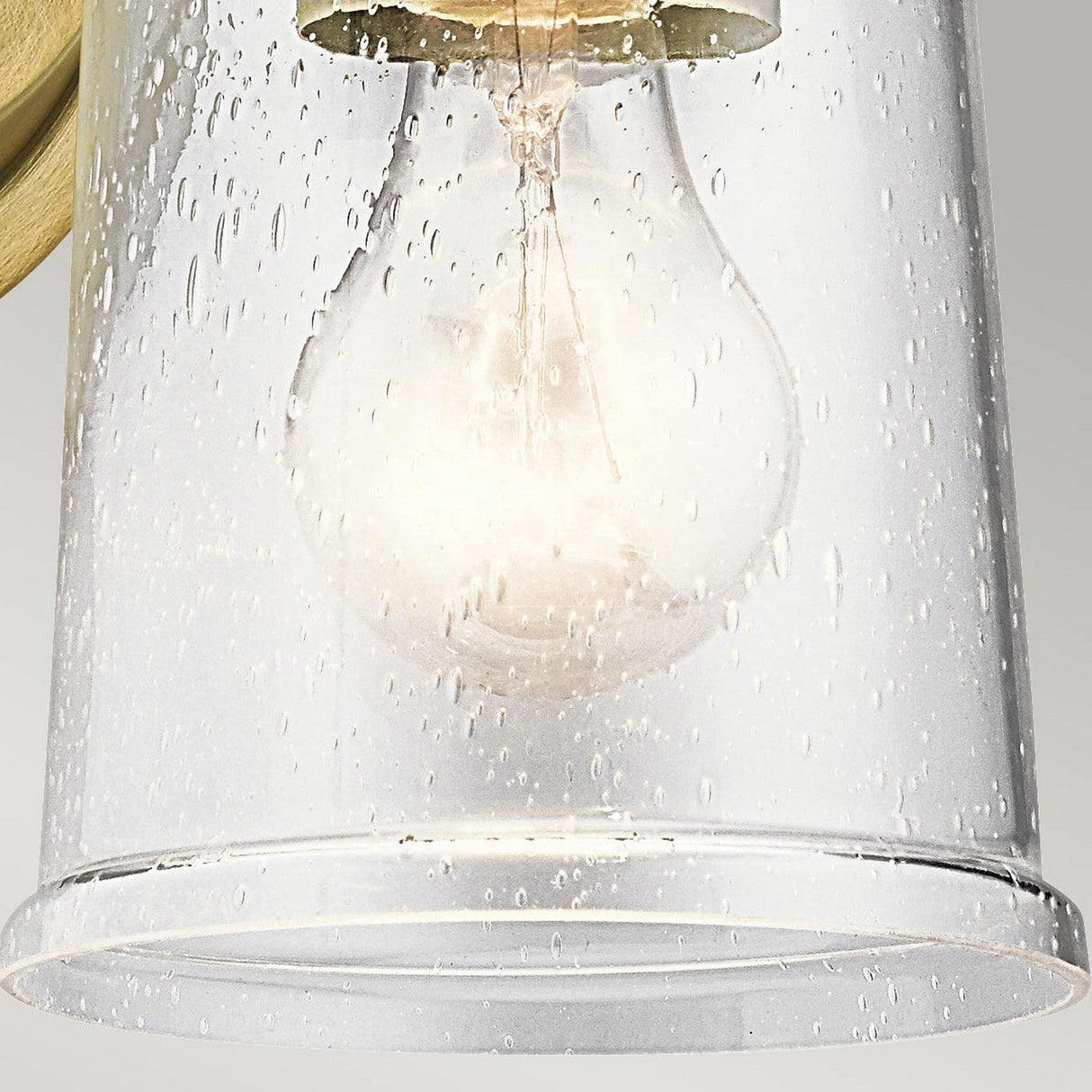 The Waverly 1 Light Wall Light - Natural Brass features a light bulb enclosed in a clear seeded glass shade with small droplets decorating the surface, creating a scattered light effect that adds vintage charm to any space.