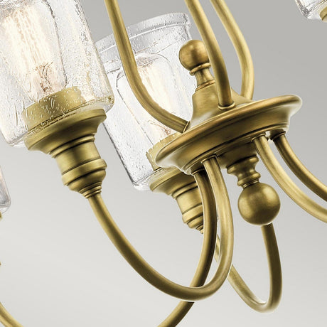 Close-up of the Waverly 5 Light Chandelier in natural brass: featuring multiple arms and seeded glass shades that reveal visible light bulbs. The sleek, curved metallic design creates a striking contrast with the textured glass, set against a light gray background.