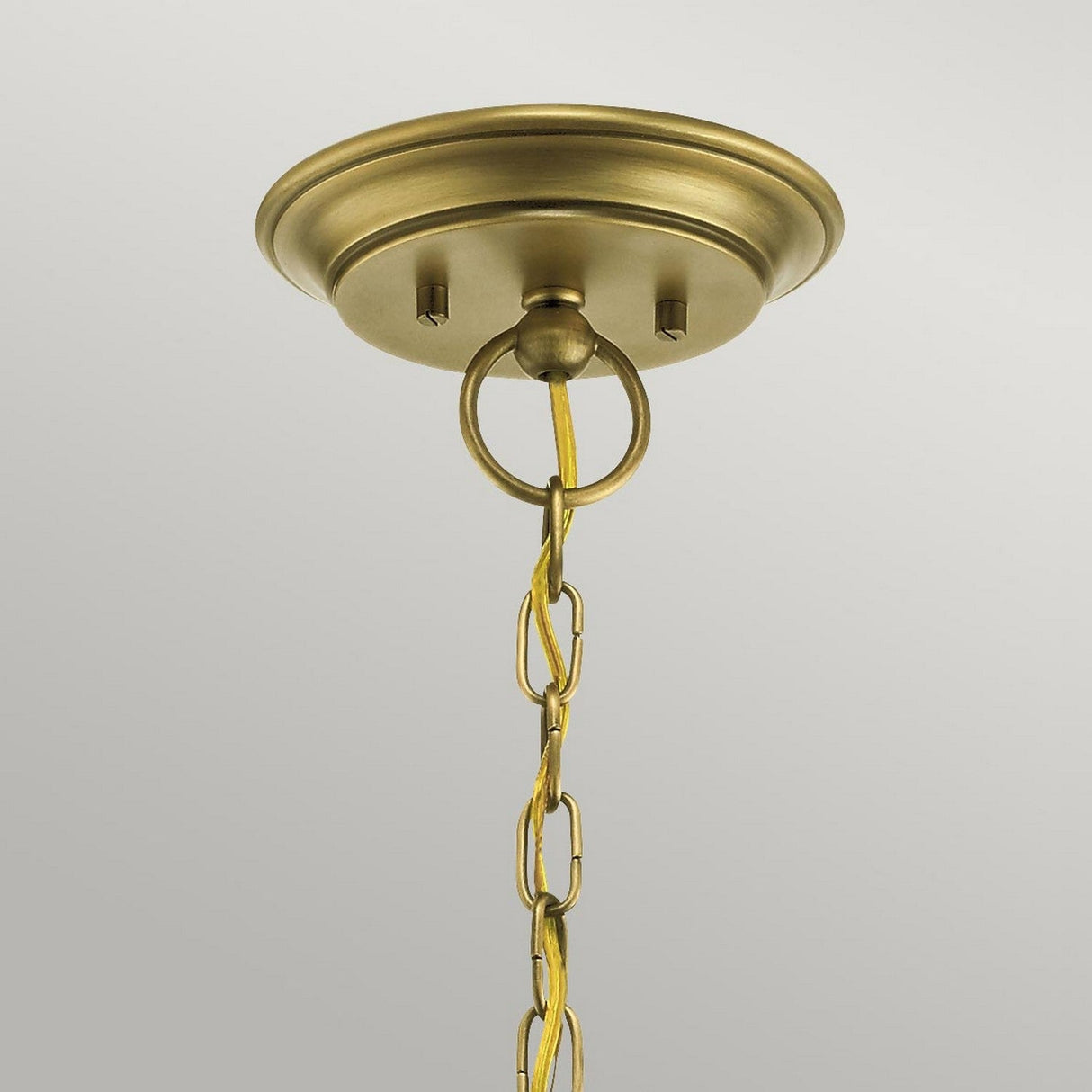 The Waverly 5 Light Chandelier in natural brass gracefully hangs down with its decorative ring and chain, which suspends the light fixture and enhances its vintage charm. The backdrop is plain and light-colored, highlighting the chandelier’s elegance.