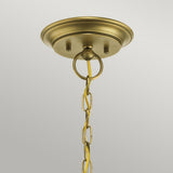 The Waverly 5 Light Chandelier in natural brass gracefully hangs down with its decorative ring and chain, which suspends the light fixture and enhances its vintage charm. The backdrop is plain and light-colored, highlighting the chandelier’s elegance.