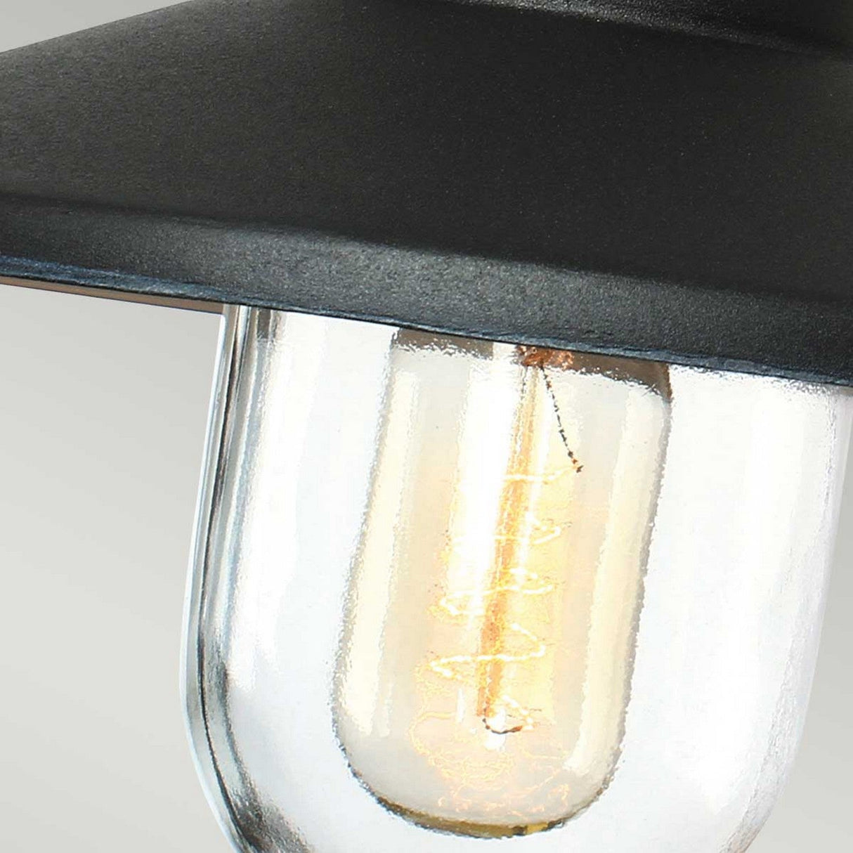 Close-up of the Klampenborg 1 Light Outdoor Wall Light in black, featuring a vintage-style bulb embraced by a clear glass casing and partially covered by a black metallic shade. The warm glow of the filament captures Scandinavian design principles, making it ideal for an outdoor wall light setting.
