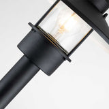 A detailed view of the Klampenborg 1 Light Bollard in black features a clear glass enclosure with an illuminated bulb. The contemporary, minimalistic design enhances the outdoor lighting aesthetic against a simple white background.
