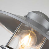 Close-up of the Klampenborg 1 Light Bollard in silver, featuring a clear glass casing that reveals a glowing incandescent bulb. This corrosion-resistant fixture boasts a domed top and a wide, round shade, all set against a plain background, embodying a touch of Scandinavian design.