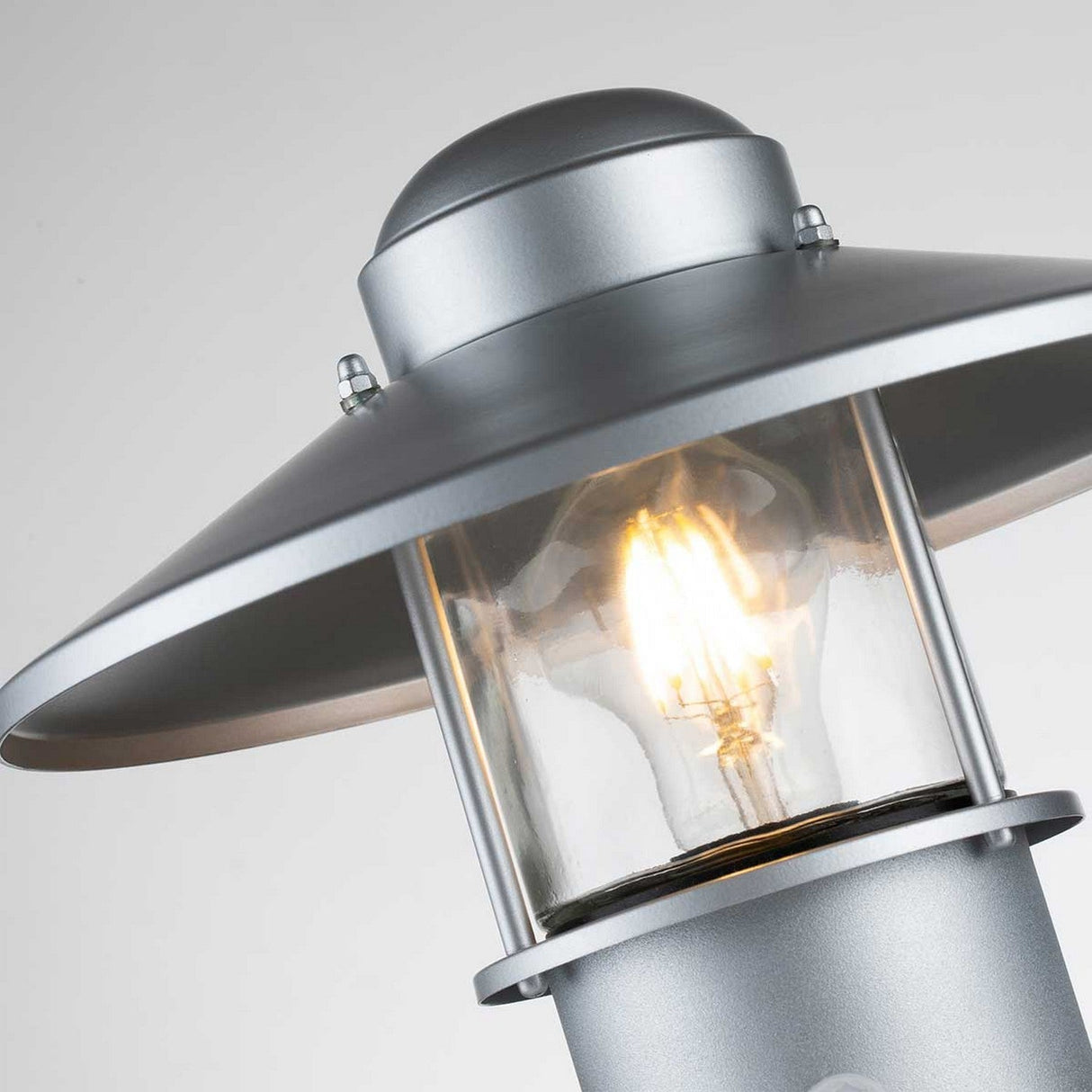 Close-up of the Klampenborg 1 Light Pedestal with PIR in silver, highlighting its modern Scandinavian design with a clear glass cover that reveals an illuminated bulb inside. The lamp features a conical top and is set against a neutral background, exemplifying sleek outdoor lighting.