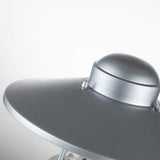 A close-up view of the Klampenborg 1 Light Pedestal in silver reveals its metallic-gray, dome-shaped design with a smooth, reflective surface. This contemporary outdoor light fixture is displayed from a slight angle, showcasing a few bolts near the top against a plain light gray background that highlights its sleek design.