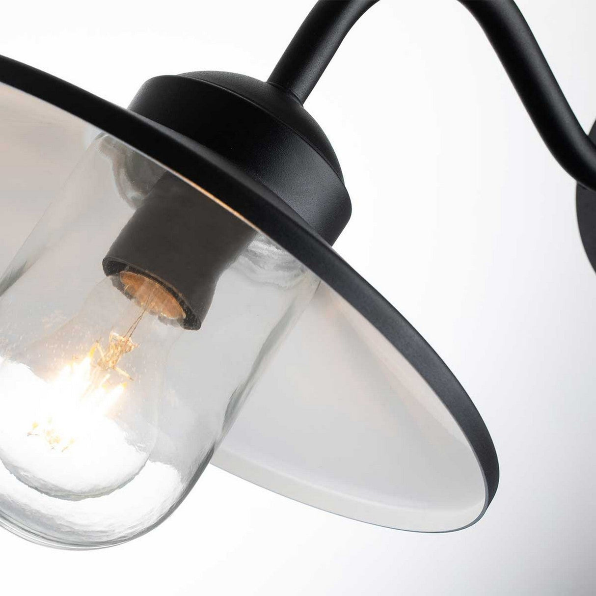 A close-up view of the illuminated Klampenborg 1 Light Outdoor Wall Light with PIR showcases its clear glass fixture and wide, white metal shade. The lamp's black metal frame beautifully merges modern industrial style with Scandinavian design, while the softly blurred background enhances its sleek aesthetic.