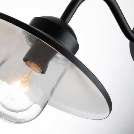 A close-up view of the illuminated Klampenborg 1 Light Outdoor Wall Light with PIR showcases its clear glass fixture and wide, white metal shade. The lamp's black metal frame beautifully merges modern industrial style with Scandinavian design, while the softly blurred background enhances its sleek aesthetic.