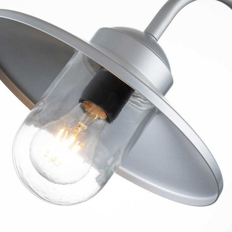 A close-up image showcases the Klampenborg 1 Light Outdoor Wall Light with PIR - Silver, featuring a glowing bulb encased in a clear glass cover. It is attached to a sleek stainless steel fixture with a wide, silver reflective shade and emits a warm glow against the white background.