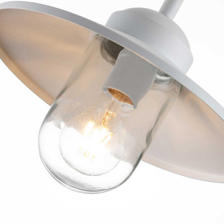 Close-up of the Klampenborg 1 Light Outdoor Wall Light in white, featuring a clear glass bulb and a wide, white metal shade. The bulb emits a warm glow that enhances its minimalist, industrial design. Reminiscent of Scandinavian outdoor wall lights, it adds elegance against the plain white background.
