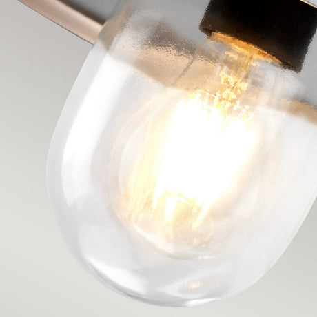 A close-up of the Klampenborg 1 Light Outdoor Wall Light - Silver, featuring a glowing light bulb inside a clear glass fixture encased in sleek stainless steel, emits a warm and bright light against a muted gray backdrop. Its reliable IP44 rating makes it perfect for outdoor wall lighting.