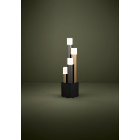 Korbelle LED table lamp with four stepped square columns in black, brown and gold, each topped with a cube-shaped satin shade, set on a black square base against a dark green background.