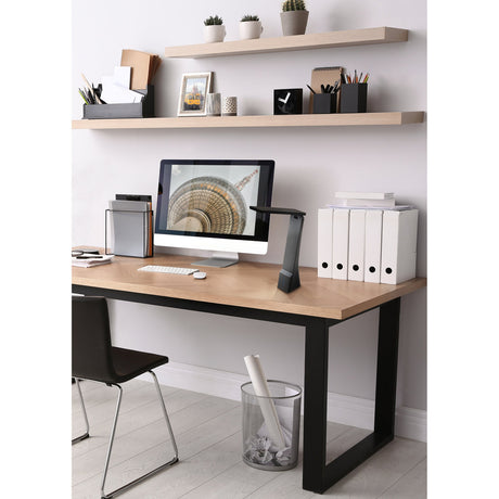 La Seca LED touch table lamp in black plastic with angled rectangular head on a tapered stem, shown on a desk beside a monitor in a home office setting.