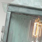 A close-up of the Lambeth Palace 1 Light Outdoor Wall Light - Verdigris reveals its weathered charm with a glowing filament against a light background. The solid brass frame showcases an aged, rustic texture, and the filament emits a warm light that beautifully contrasts with the greenish hue of this exquisite outdoor fixture.