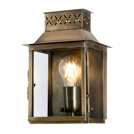 The Lambeth Palace 1 Light Outdoor Wall Light - Aged Brass boasts a glass enclosure with a visible glowing bulb, radiating vintage charm. Its aged brass finish and ornate cut-out patterns on the top enhance elegance in any outdoor environment.