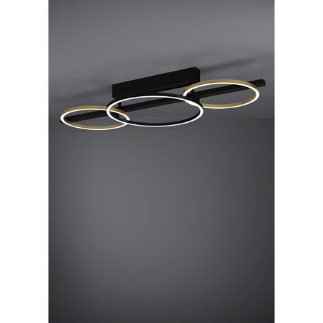 Lanacera LED ceiling light with three interlinked rings on a black bar, gold rings at each end and a black ring at the centre, lit against a dark grey background.