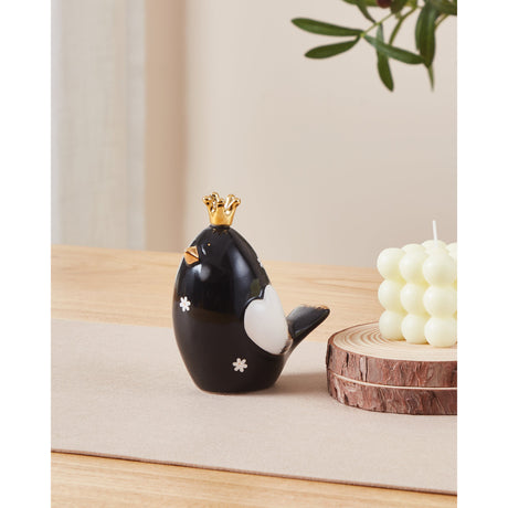 Landjut ceramic bird ornament with glossy black body, white wing detail, small white flower accents and a gold crown and beak, shown on a wooden table beside a candle.
