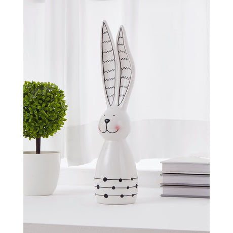 White and black ceramic rabbit ornament with tall zigzag ears, a smiling dotted face, and dotted bands on a bell-shaped body, displayed on a shelf beside books and a potted plant.