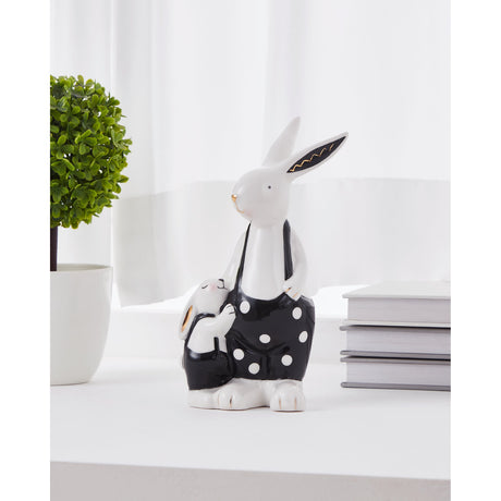Landjut ceramic rabbit ornament in white and black with gold accents, tall rabbit in polka-dot dungarees beside a smaller nestled rabbit, shown on a shelf near books and a plant.