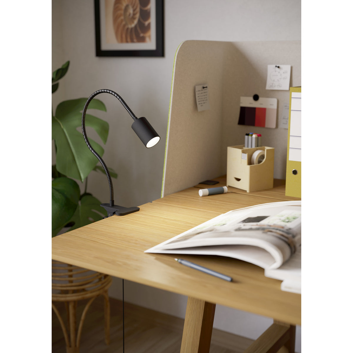 Lardaro Clamp Lamp in black with flexible gooseneck arm and cylindrical head, clipped to a desk edge in a home study beside a plant and noticeboard.