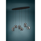 Black Larva linear pendant light with six staggered smoked glass barrel and globe shades on a long oval ceiling plate, shown against a dark teal background.