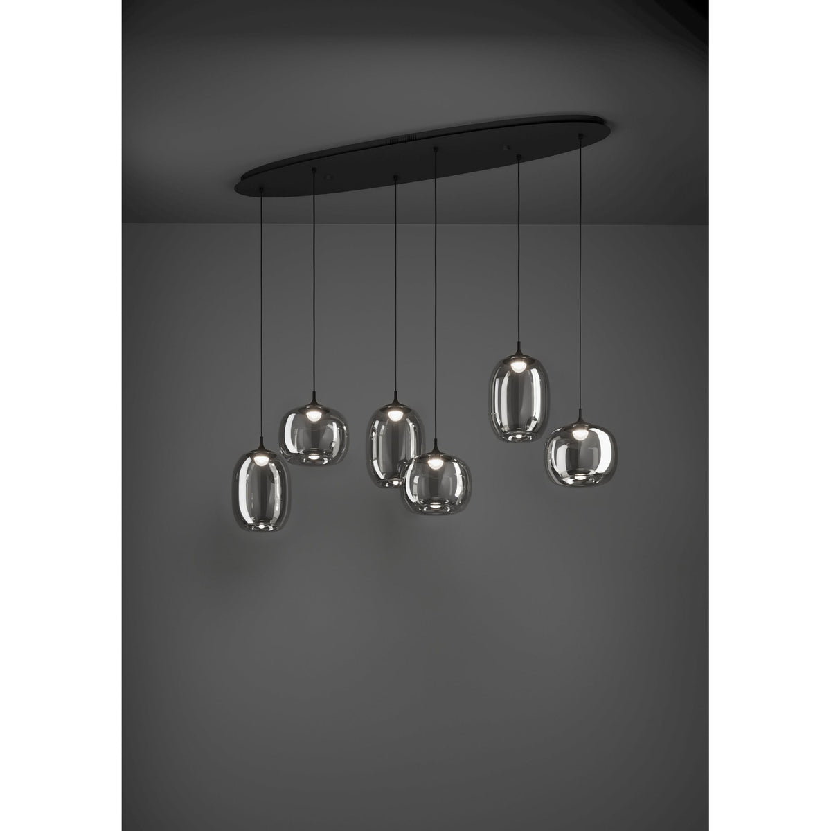 Larva Linear Pendant Light in black steel with six smoked glass shades of staggered barrel and globe shapes, suspended from a long oval ceiling plate against a dark background.