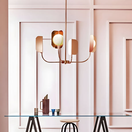 A modern interior features the Petal 4 Light Chandelier in copper, with its geometric design hanging above a sleek glass table. The table is decorated with items like a brown pitcher and colorful cups, set against an elegant paneled wall where soft lighting enhances the contemporary decor.