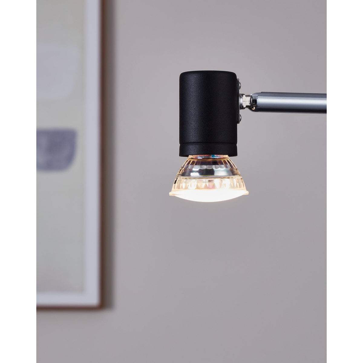 LED Bulb GU10 Warm White fitted into a black cylindrical spotlight head on an adjustable arm, glowing warm white with a faceted glass body and frosted lens, against a blurred interior background.