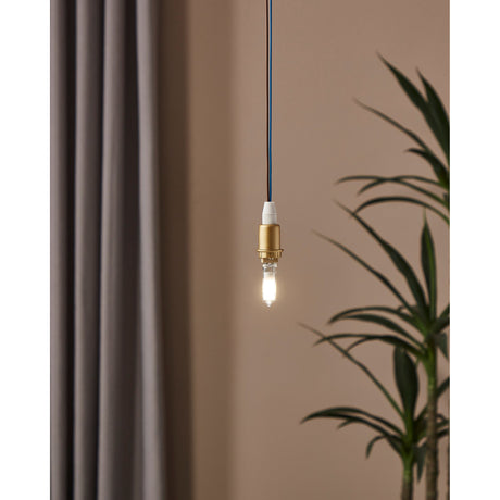 Pendant light fitted with a G9 LED capsule bulb, brass holder and blue cable, glowing warm against a muted pink wall with curtain and plant nearby.