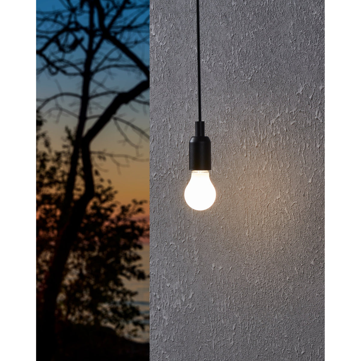 Pendant light with a black cable and fitting holding an LED GLS E27 bulb, glowing warm white against a textured grey wall at dusk.