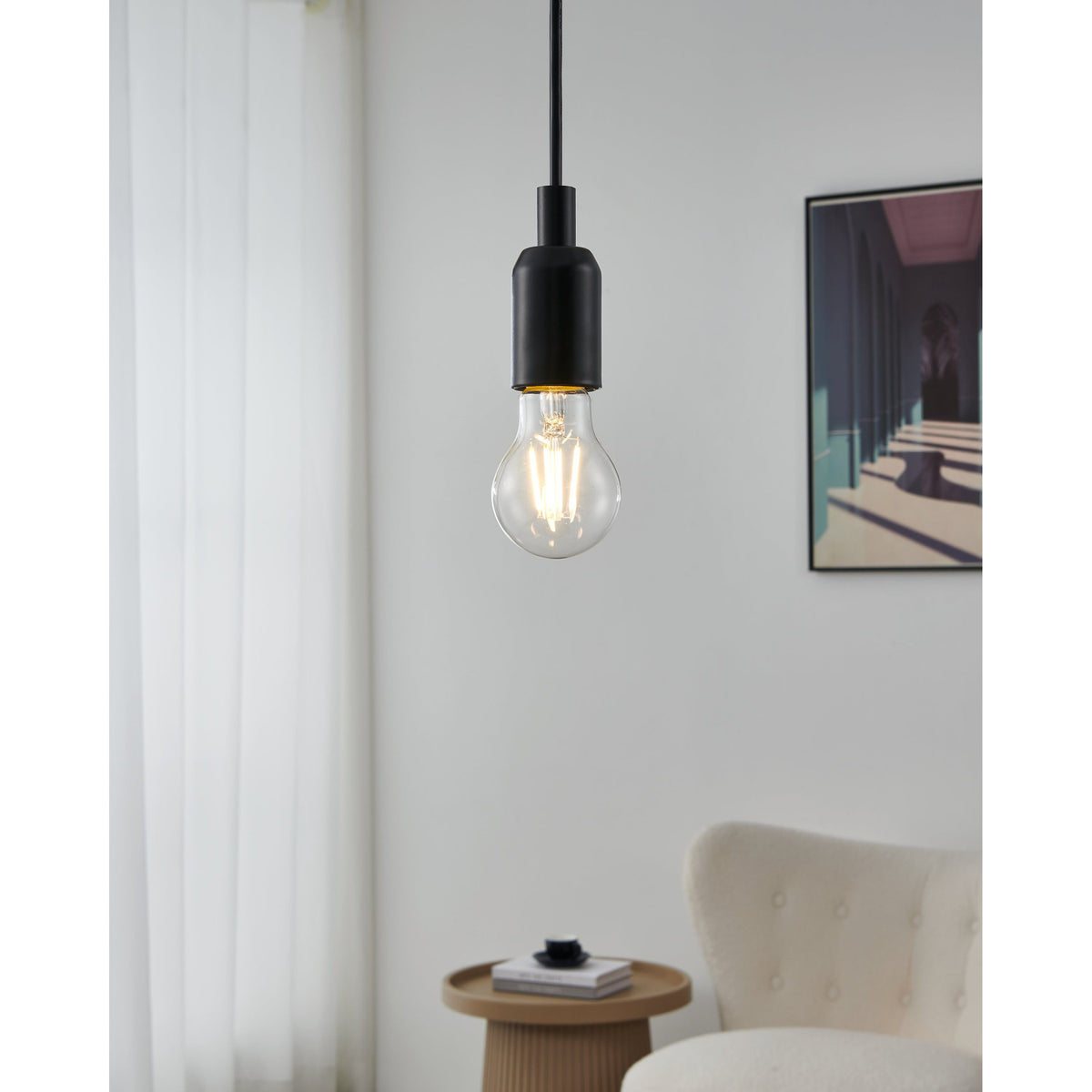 Pendant light with black fitting and clear glass LED GLS bulb showing visible filament strips, hanging in a living room near a curtain, side table, armchair and framed artwork.