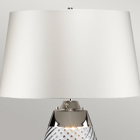 The Lena 2 Light Small Blue Table Lamp showcases an off-white fabric shade paired with a textured, mouth-blown glass base. It is accentuated by a polished metal detail connecting the base to the shade, all highlighted against a neutral gray backdrop.
