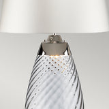 The Lena 2 Light Small Blue Table Lamp features a twisted, mouth-blown glass base with an elegant metal accent, all complemented by an off-white shade. This stylish table lamp offers a modern look while softly illuminating the intricate design of the glass.