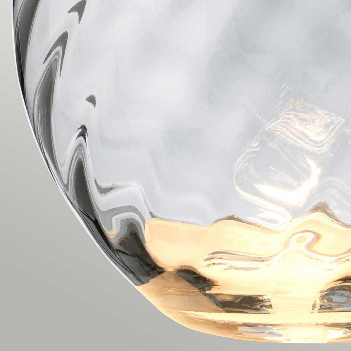 A close-up view of a smoke-hued glass piece showcases wavy textures with a transparent surface. The lower section transitions from golden to clear, capturing and reflecting light in an abstract, artistic design akin to the sophisticated style of the Lena 2 Light Large Smoke Table Lamp with Slate Shade. The background is softly colored in neutral tones.